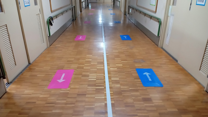 廊下の片側に青の誘導マット、もう片側にピンクの誘導マットが等間隔で設置。それぞれ奥へ向かう矢印、手間に向かってくる矢印が表示されている。