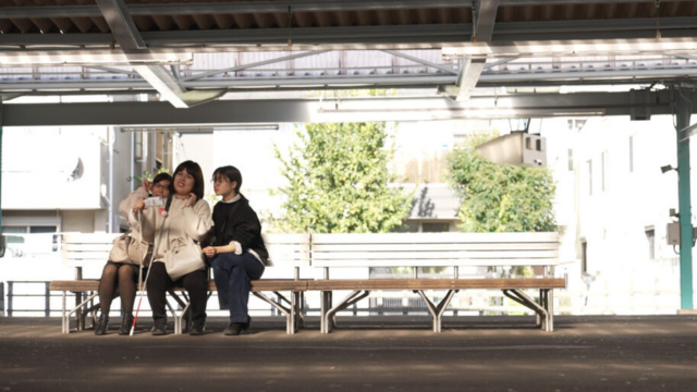 電車のホームベンチで座る女性三人組。一人は白状を持っている