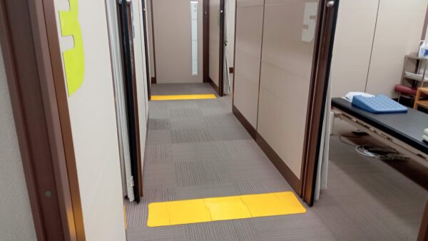 タイルカーペット貼りの廊下で、長手方向とは垂直に誘導マットが２列設置されている。設置されているのはマッサージルームの入口で、部屋の入口位置が誘導マットでわかるようになっている。