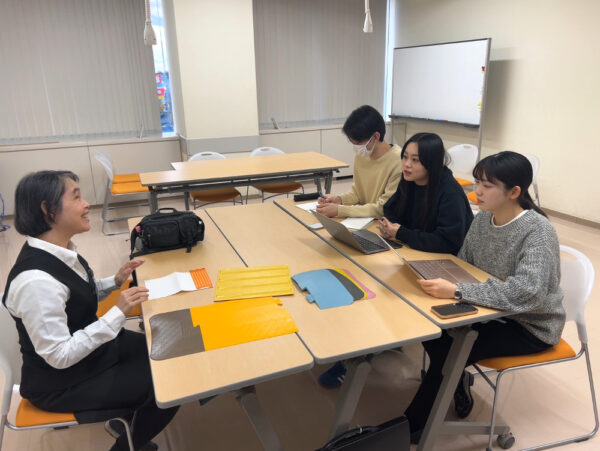 テーブルの対面に座り、笑顔で会話する亀山様とインタビュアー３人を、側面から撮影した写真。