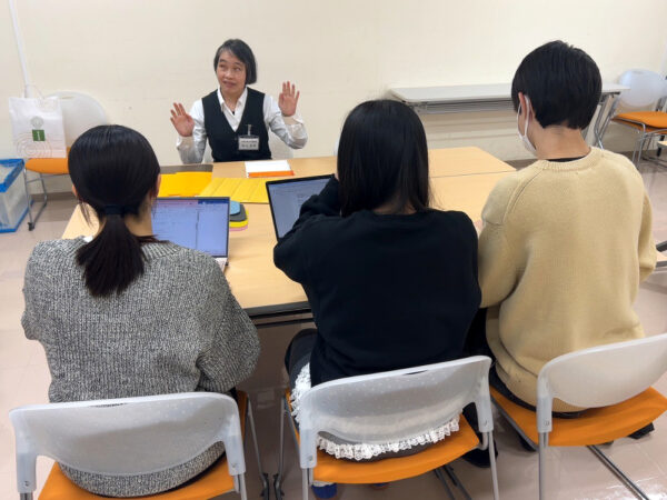 テーブルの対面に座り、笑顔で会話する亀山様とインタビュアー３人を、インタビュアーの後ろから撮影した写真。