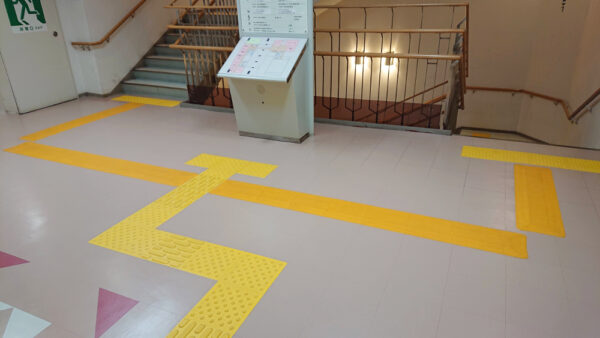 階段の踊り場の広い空間に誘導マットが案内板を避けるようにコの字に設置されている写真。