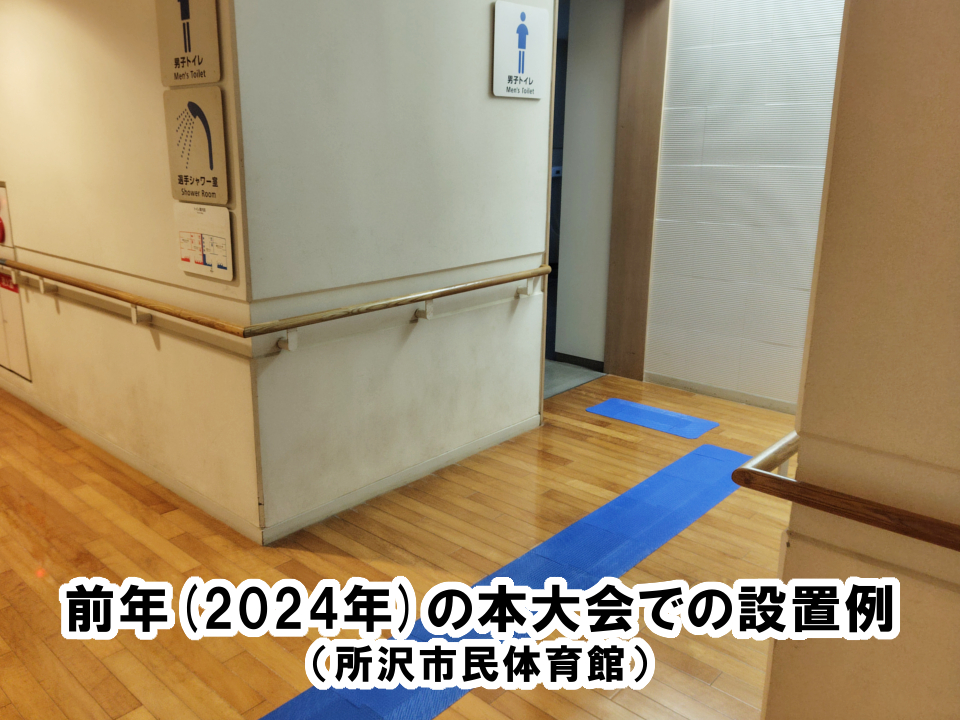 男子トイレ入口に向かって、L字型にブルーの誘導マットが設置されている写真。下部にテキスト→前年(2024年)の本大会での設置例　（ 所沢市民体育館）